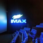 An IMAX movie theater auditorium with blue seats and the IMAX logo glowing beside the screen